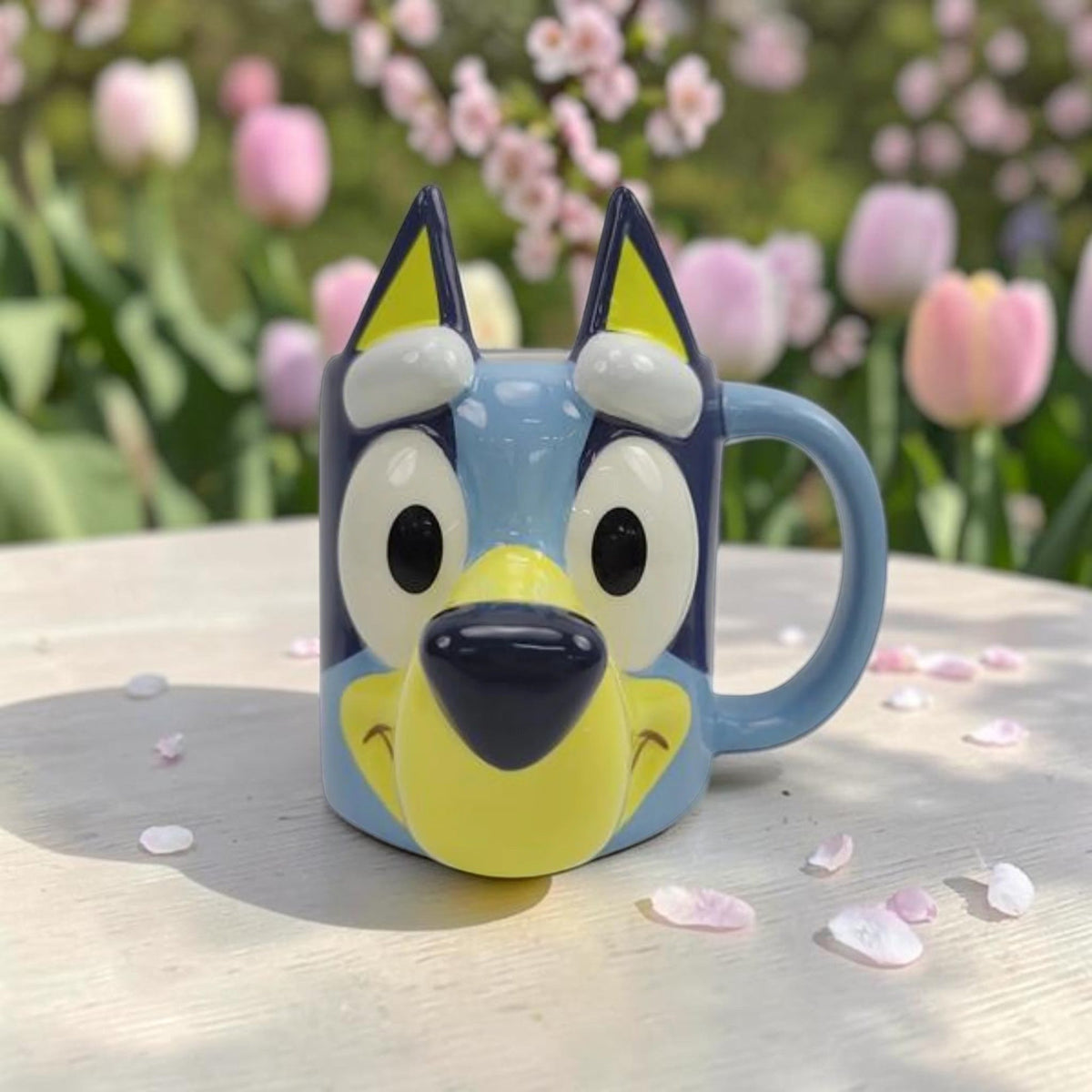 Taza 3D de cerámica Magia Nostálgica con forma de la cara de Bluey en tonos azules y amarillos sobre mesa de jardín